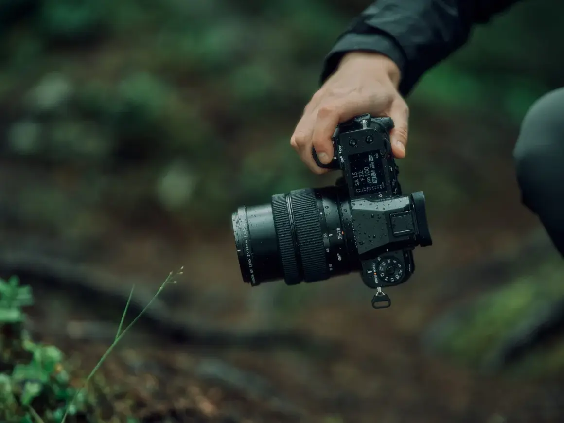 En hand håller en regnvåt Fujifilm GFX 50S II med ett zoomobjektiv i skogen.