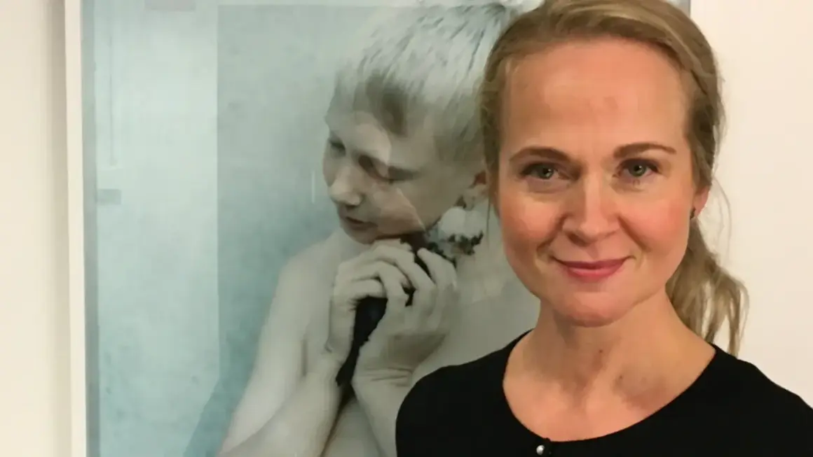 Anna Claren ler mot kameran, med en konstnärlig bild av en pojke som håller en fågel i bakgrunden.
