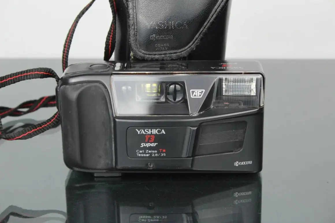 Svart kompaktkamera, en Yashica T3 Super med Carl Zeiss Tessar-objektiv, med en svart läderfodral och en röd-svart kamerarem.