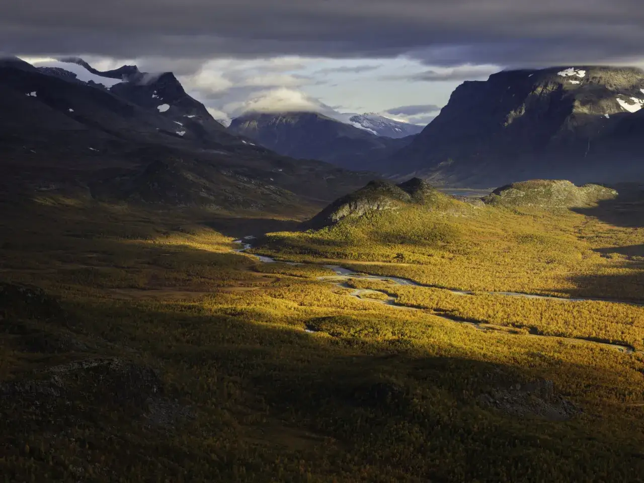 Magnus Lindbom fångar höstens gyllene ljus över fjäll och en slingrande älv. Dramatiska bergskedjor reser sig mot en molnig himmel.