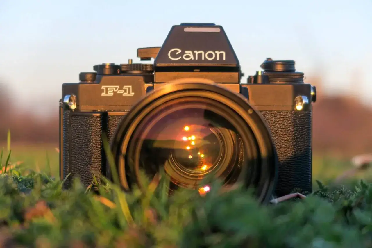 En svart Canon F-1 kamera med objektivet framåt, placerad i gräset. Solnedgångens reflektioner syns i linsen.