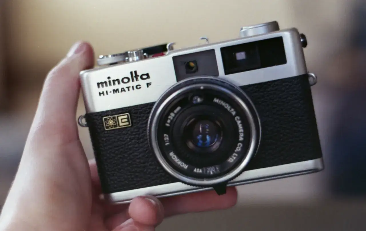 En hand håller en silverfärgad Minolta Hi-Matic F-kamera med svart läderdetalj.