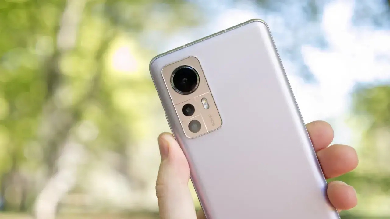 En hand håller en ljuslila Xiaomi 12 för ett test. Kameramodulen med 50MP syns tydligt mot en suddig grön bakgrund.