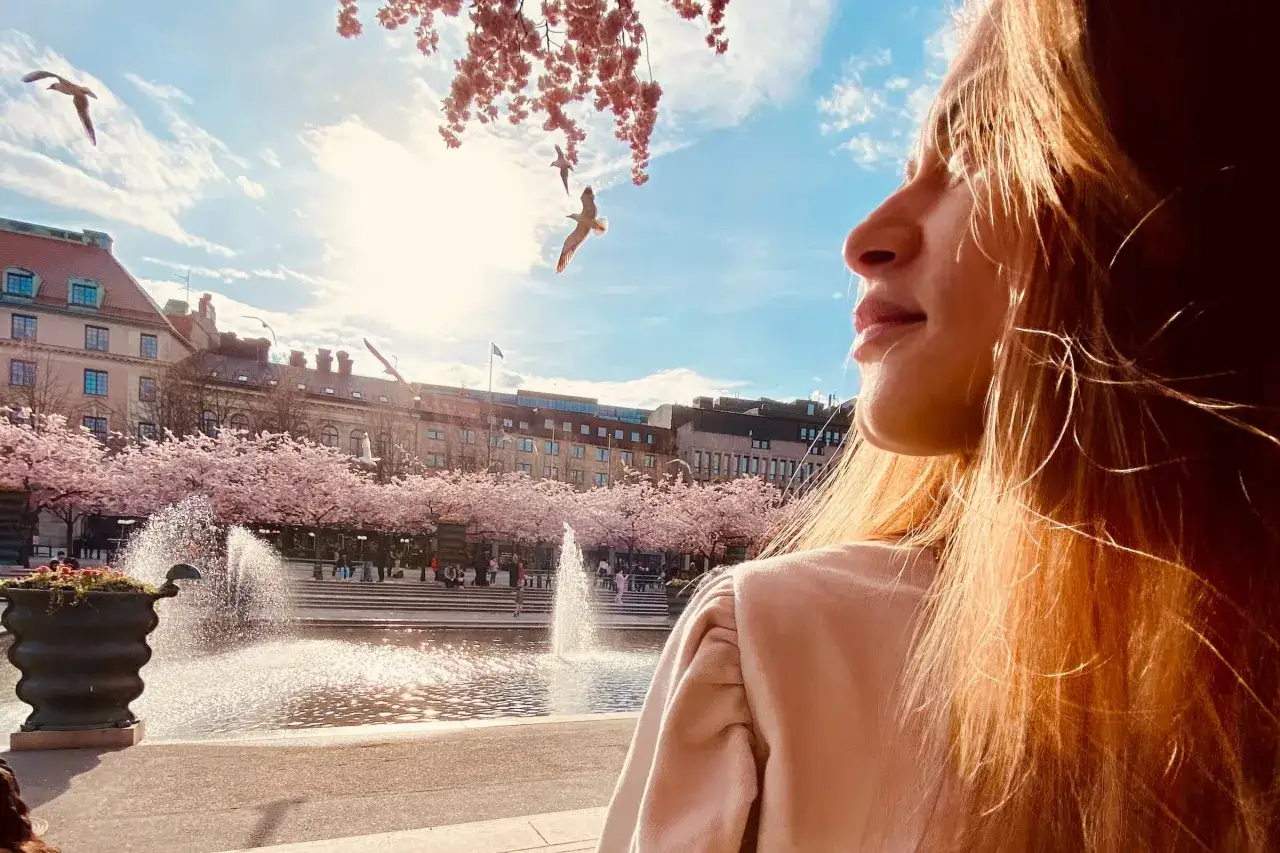 En ung kvinna njuter av v&aring;ren vid en font&auml;n i Stockholm. K&ouml;rsb&auml;rstr&auml;den blommar och m&aring;sar flyger &ouml;ver himlen.