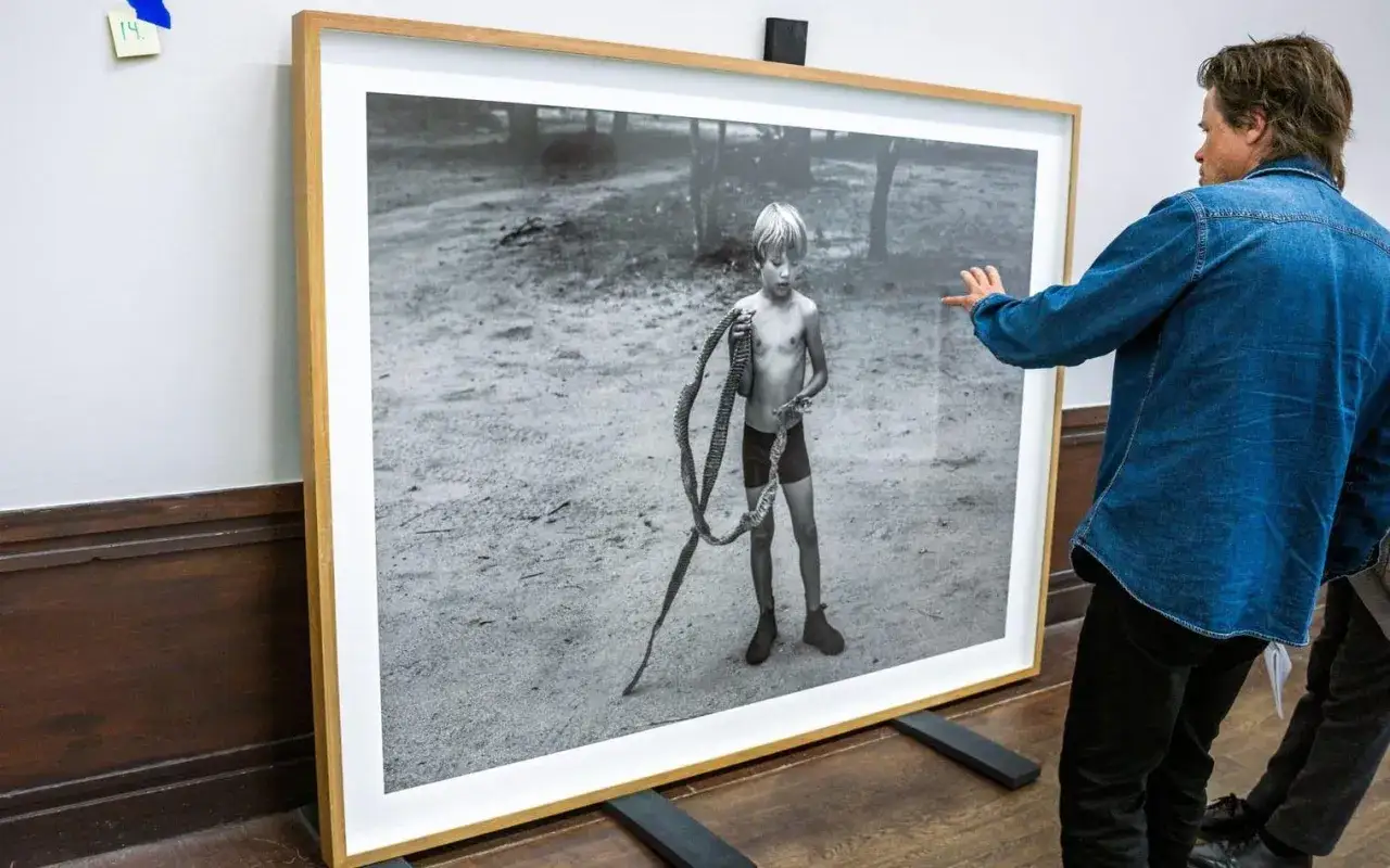 En man i jeansjacka pekar på ett stort svartvitt fotografi av en pojke med ett rep, troligen ett verk av Jens Assur.