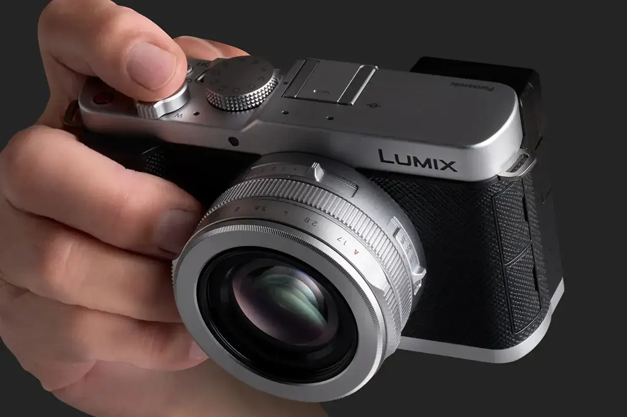 En hand håller en silverfärgad Panasonic LX100-kamera med en objektivring som visar bländartal.
