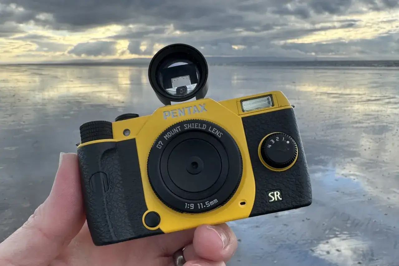 En gul och svart Pentax Q-kamera hålls upp mot en molnig himmel och en spegelblank strand.