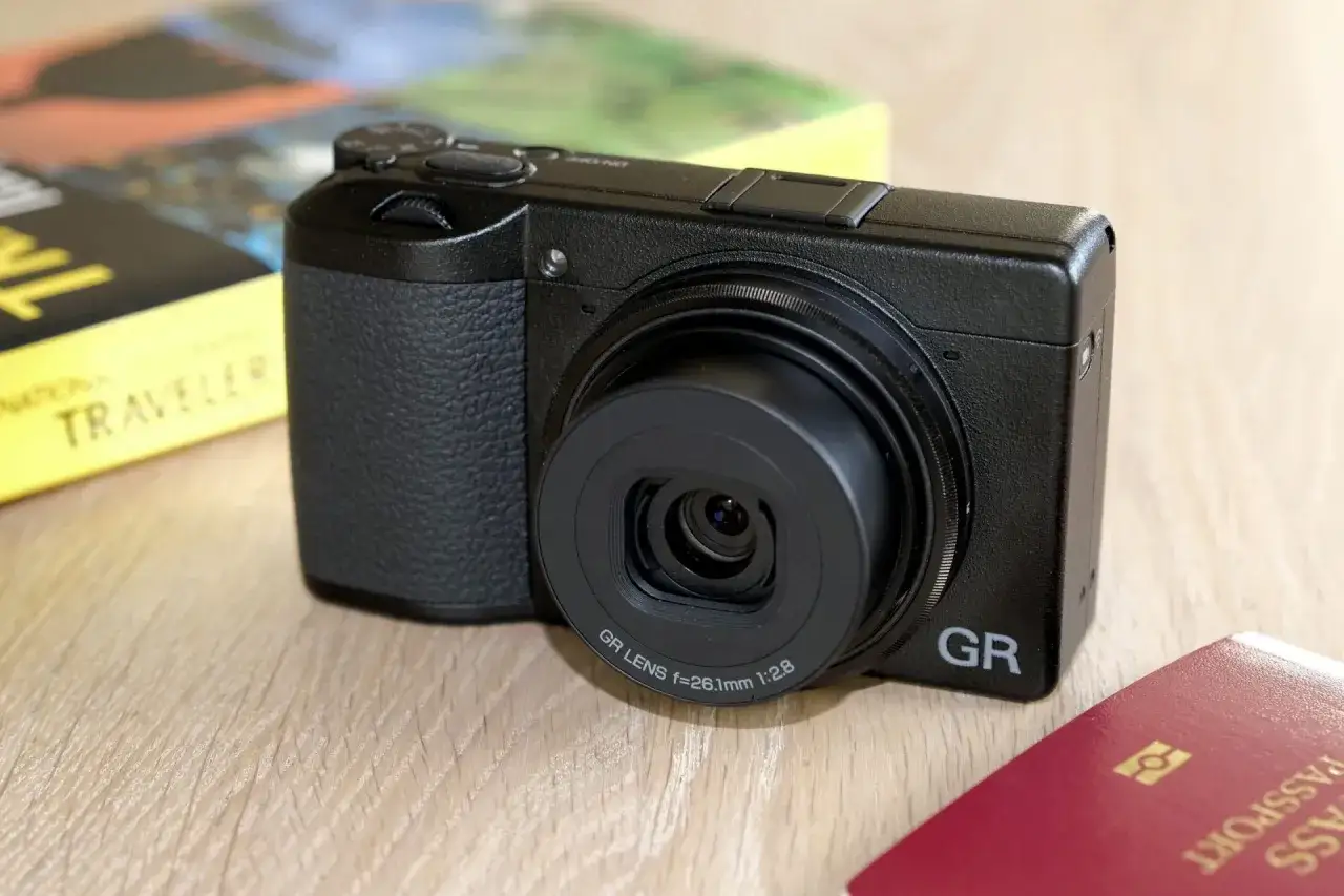 En svart Ricoh GR III X-kamera ligger på ett träbord bredvid en resebok och ett pass. Perfekt för att fånga dina äventyr.