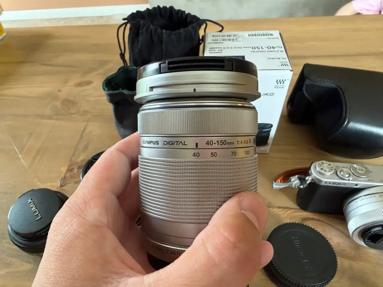 En hand h&aring;ller ett silverf&auml;rgat Olympus-objektiv, redo f&ouml;r en Panasonic GM1. Tillbeh&ouml;r som objektivlock och fodral syns.