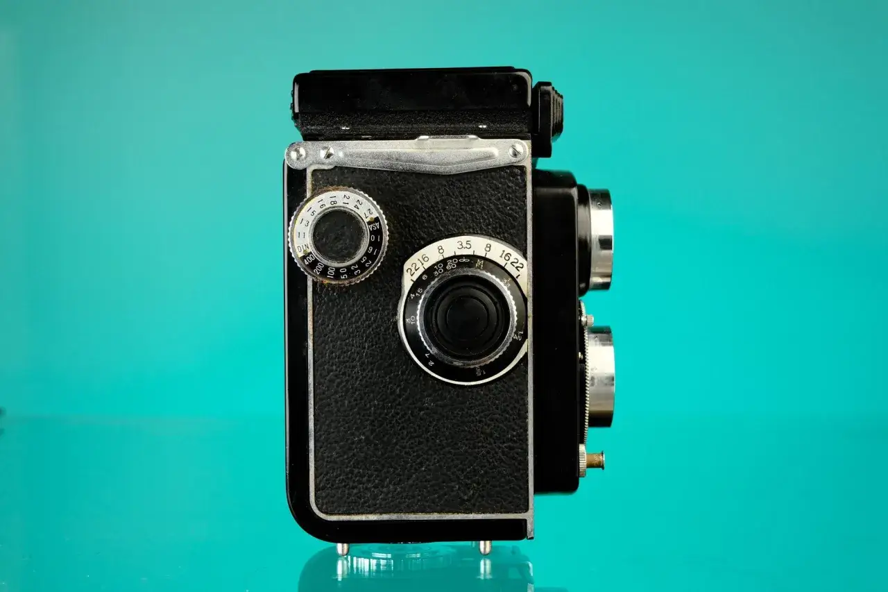 En svart, klassisk Yashica A spegelreflexkamera med kromdetaljer st&aring;r mot en turkos bakgrund.