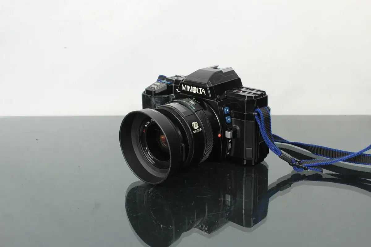 Svart Minolta 7000 systemkamera med objektiv och blå axelrem, placerad på en reflekterande yta.