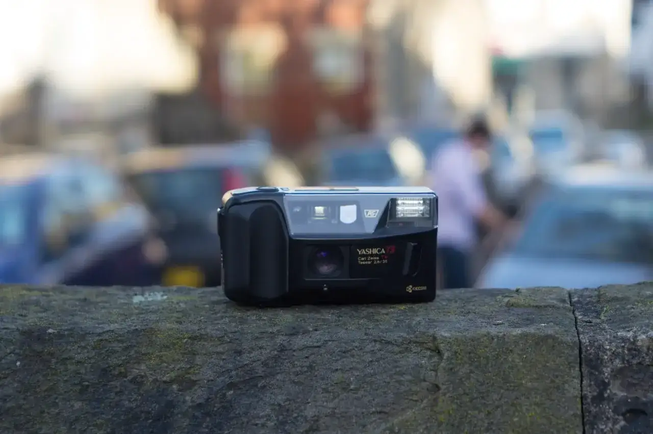 En svart Yashica T3-kamera vilar p&aring; en mossig stenmur. Bakgrunden &auml;r suddig med bilar och en person.