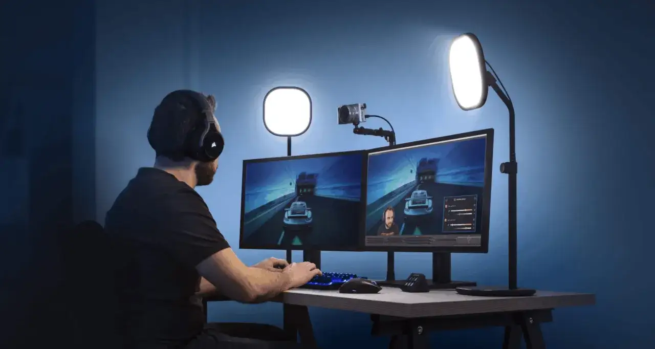 Person med hörlurar spelar datorspel, med en kamera på datorn och två skärmar som visar spelet.