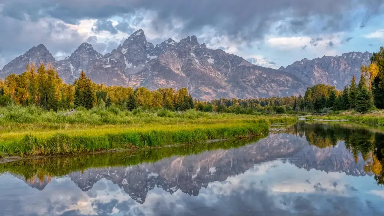 Majest&auml;tiska bergssiluetter speglas i en lugn flod, omgivna av frodig gr&ouml;nska och tr&auml;d. En vacker vy med high dynamic range f&aring;ngar detaljer i b&aring;de ljusa och m&ouml;rka partier.