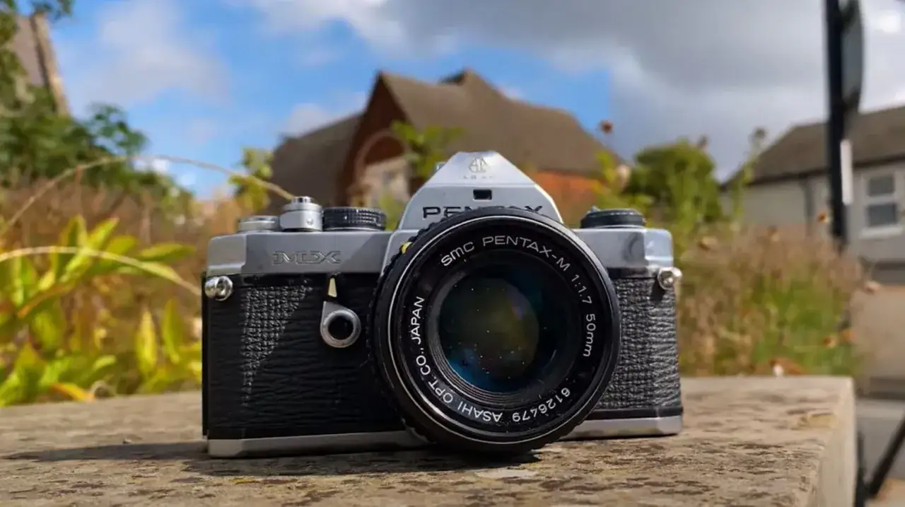 En klassisk Pentax MX-kamera med objektivet SMC Pentax-M 50mm 1:1.7. Kameran vilar på en stenmur med hus och träd i bakgrunden.