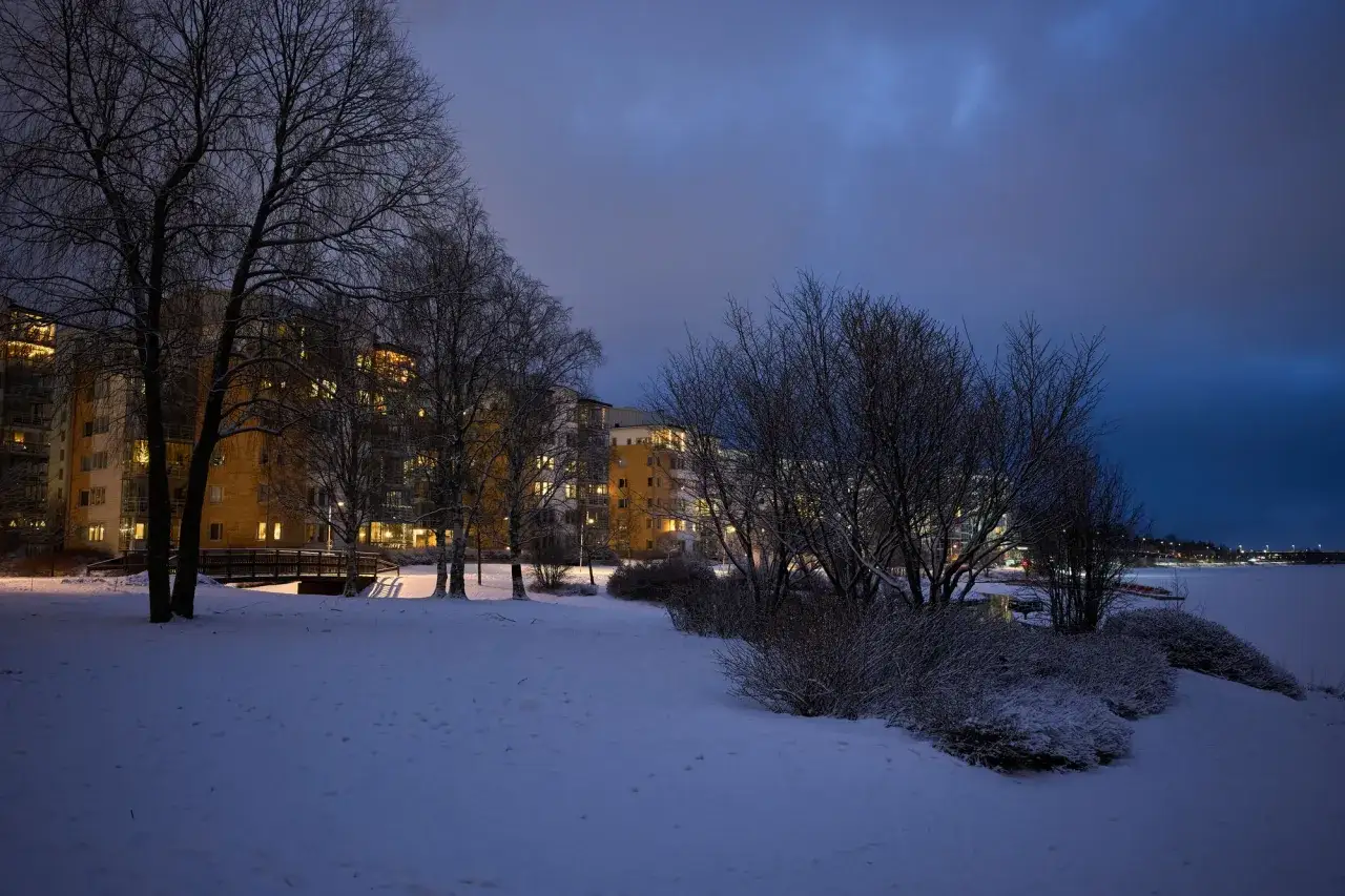 Vinterkväll i Sverige med snö, träd och upplysta lägenhetshus vid en frusen sjö. Fångat med en Ricoh GR IV.