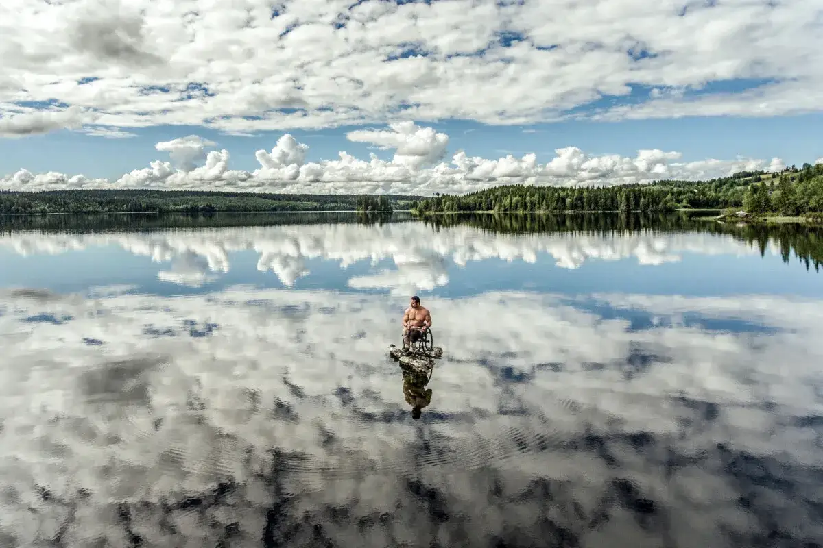 En man i rullstol sitter på en sten ute på en spegelblank sjö. Solen lyser på den molniga himlen. Sophie Odelberg fångar en stund av lugn.