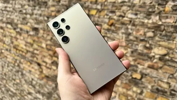 En hand håller en ljusgrå Samsung S24 med flera kameror mot en tegelvägg.