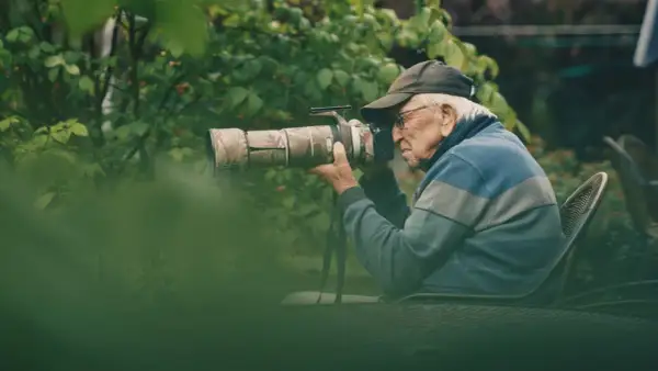 Äldre man med keps och glasögon fotograferar med en stor kamera i en lummig trädgård. En passionerad naturfotograf i sitt esse.