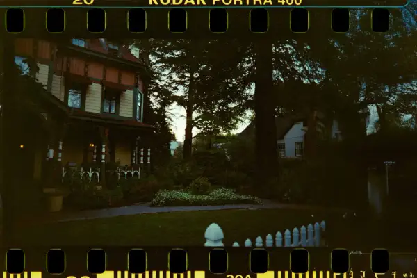 Ett gammalt hus med veranda, omgivet av träd och en trädgård. Bilden har en tydlig film format-känsla med perforeringar.