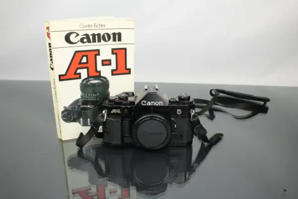 Svart Canon A-1 systemkamera med objektiv och en bok om kameran. En klassiker för den som vill lära sig mer om analog fotografering.