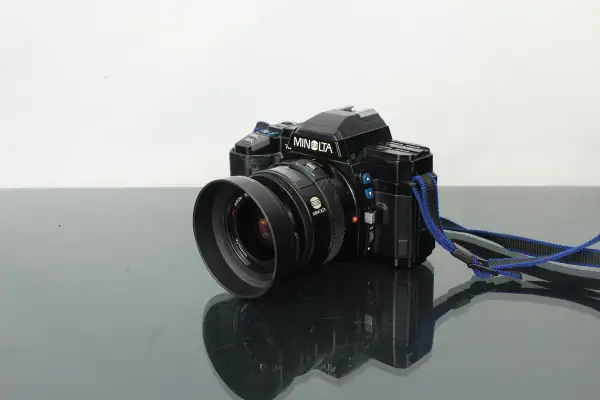 Svart Minolta 7000 systemkamera med objektiv och blå axelrem, placerad på en reflekterande yta.